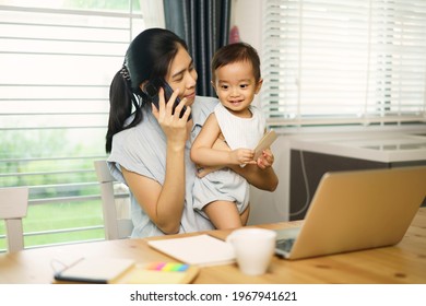 Work From Home. Asian Young Mother Calling Mobile, Working And Holding Her Son. Entrepreneur Woman Working With Laptop. A Boy Sitting With Mom At Home In Kitchen. Quarantine On Coronavirus Pandemic.