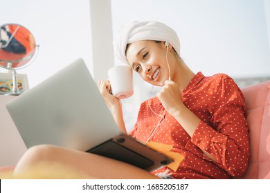 Work From Home Concept - Beautiful Young Woman In Red Pajamas Shirt With Towel Bath Head Wrap Working On Laptop, Sitting On A Couch, Holdign A Cup Of Coffe. Working At Home. Quarantine.