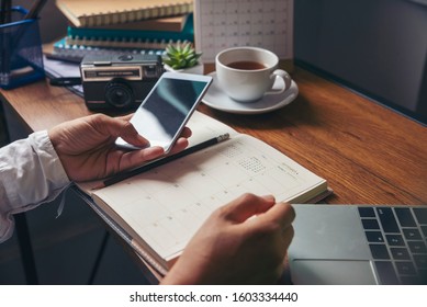 Work from home concept. Planner using phone and laptop to plan daily agenda on calendar book. Woman Hand mark and note schedule (holiday trip) on diary at business office desk. Coffee place on table