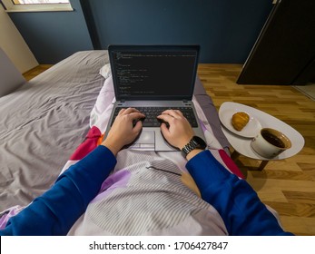 Work From Home Concept. POV View Of A Man Sitting On A Bed With A Laptop And Breakfast On A Small Table Beside The Bed.