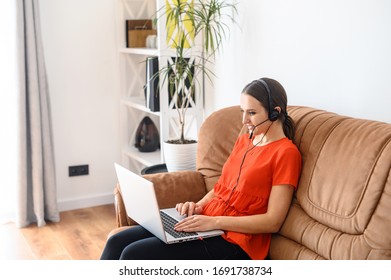 Work At Home, Distant Work, Remote Work. A Young Woman Sits On A Sofa With Hands Free Headset And Laptop, She Types On The Keyboard