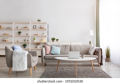 Work From Home During Covid-19 Pandemic, Renting Apartment, Buying, Ad Of Real Estate. Gray Sofa With Pillows, Table With Laptop And Cup Of Hot Drink, Shelves And Armchair In Living Room Interior