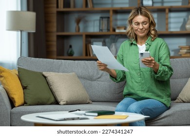 Work from home. An elderly woman in glasses is sitting on the sofa at home, holding a phone and documents in her hands. Works with papers, checks and pays online bills. - Powered by Shutterstock