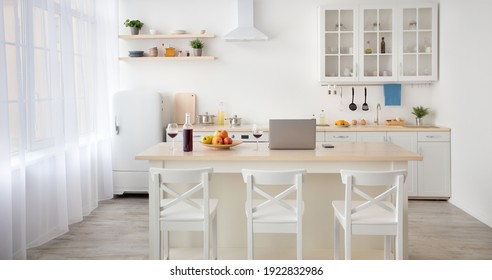 Work from home, freelance, interior blog and simple design. Laptop, fruits and wine on table, white furniture with utensils, shelves with dishes and plants in pot, refrigerator in the dining room - Powered by Shutterstock