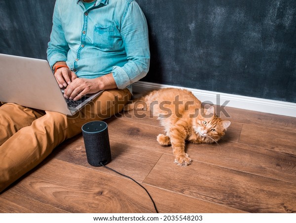 Work from home\
with funny lazy red cat pet. Man sitting on laminate wooden floor\
with laptop computer and smart speaker alexa. Ginger pet cat lying.\
Home office. Remote work