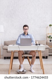 Work From Home And Humor. Busy, Serious Guy In Shirt And Shorts Works At Laptop And Puts Foot On Dumbbell In Interior Of Living Room, Free Space