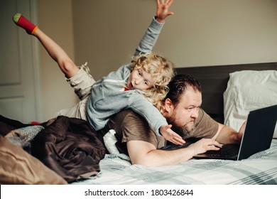 Work from home with kids children. Father working on laptop in bedroom with child daughter on his back. Funny candid family moment. New normal during coronavirus self-isolation quarantine.
