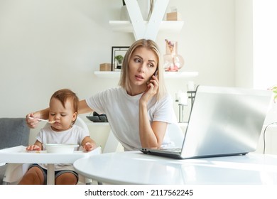 Work from home. Young mother is talking on phone while sitting at table with a laptop and feeding her little daughter. Motherhood, maternity, childhood and care concept. - Shutterstock ID 2117562245
