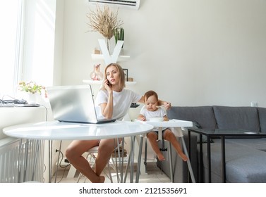 Work from home. Young mother is talking on phone while sitting at table with a laptop and feeding her little daughter. Motherhood, maternity, childhood and care concept. - Shutterstock ID 2121535691