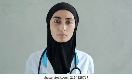 Work In A Hospital For Women Of Islam. An Educated Female Doctor Of Arab Appearance Looks Into The Camera. Portrait Of A Muslim Woman Doctor In A Medical Gown And Black Hijab.