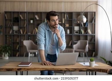 Work on idea. Thoughtful casual male in glasses stand by desk with pc look at distance touch chin create project ponder on business plan. Pensive young man scientist do research think on new solution