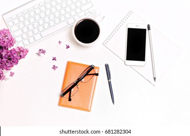 Work place with computer keyboard, mobile phone, cup of coffee, notebook, pencil, pen, glasses and lilac flowers on the white table background. Flat lay, top view