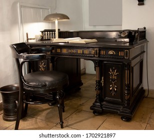 A work place with a vintage, dark brown, richly ornamented, oak desk, rounded, wooden, leather armchair, telephone made of bakelite, lamp and trash can in form of mortar.