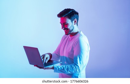 Work And Play Online. Happy Guy Holding Laptop And Typing In Neon Light, Copy Space