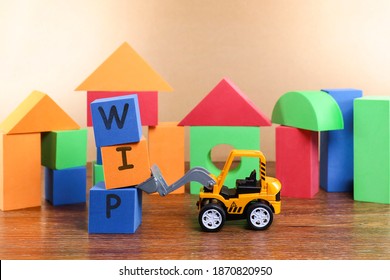 Work in Progress concept.  A toy bulldozer lifting cubes with letters on WIP and colorful blocks with various shapes at the back