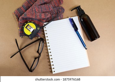 Work in progress concept.  View from above of a notebook, pen, eyeglasses, measuring tape, knitted mitted, and hand sanitizer on the brown background 