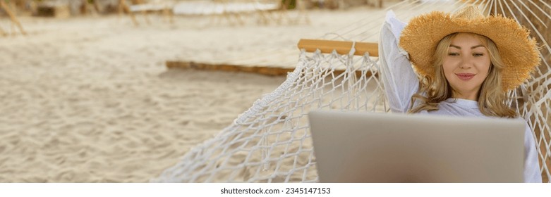 Work remotely. Side view of a young happy female freelancer in a straw hat working on a laptop while relaxing in a hammock on the beach at sunset. Remote work during vacation