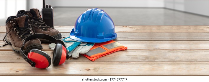 Work Safety Protection Equipment. Industrial Protective Gear On Wooden Table, Blur Office Or Home Interior Background. Hardhat, Boots, Earmuffs, Gloves, Goggles