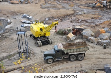 Work Site In Granite Quarry With Old Rusty Empty Truck, Yellow Crane And Lighting Tower Among The Stones