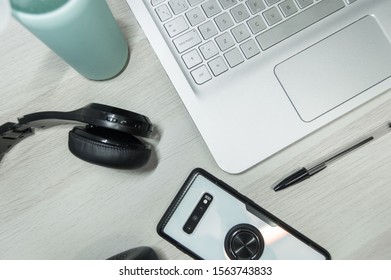 Work Student Desk With A Smartphone, Pen, Headphones, Laptop And Glass Bottle Of Water On Wooden Table.