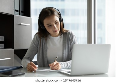 Work And Study. Interested Millennial Woman Employee Trainee Intern Sitting At Desk Wearing Wireless Headphones Watching Webinar On Computer Screen Writing Up Valuable Information On Paper Sheet