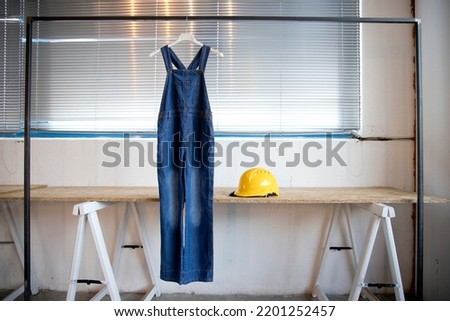 work and training, image of a worker's overalls hanging on a clothes hanger in a room with a wooden table with a yellow helmet on top