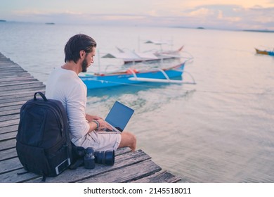 Work And Travel. Young Man With Rucksack Using Laptop Computer Sitting On Wooden Fishing Pier With Beautiful Tropical Sea View.