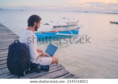 Work and travel. Young man with rucksack using laptop computer sitting on wooden fishing pier with beautiful tropical sea view.