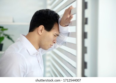 At work. Upset young brunette male standing at the window, leaning his arm and head on blinds - Shutterstock ID 1712544007