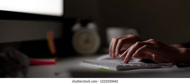 Workaholic Hand Typing Keyboard In Dark Late Night With Computer Screen Light, Clock, Coffee Cup And Stuff On Desk Table With Noir Working In Workplace Or Hacker Cyber Crime Concept. Work Hard Banner.