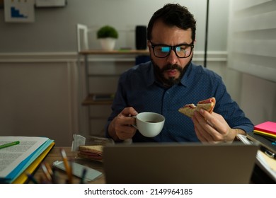 Workaholic Man With Glasses Using The Laptop While Eating A Sandwich And Drinking Coffee While Working Late 