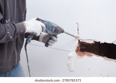 A worker aims a drill at a wall to create a hole. Dust flies as the tool breaks through the surface. The setting is a construction site with visible repairs in progress.