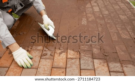 Worker applying a layer of wet mortar or grout over interlocking bricks, filling joints to create a solid patio surface