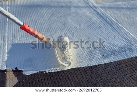 Worker applying white polyurethane waterproof coating on flat roof surface with paint roller.