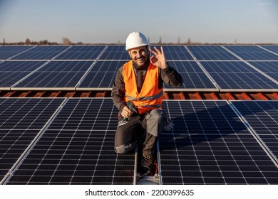 A worker is approving eco life while crouching next to the solar panels and he is smiling at the camera.