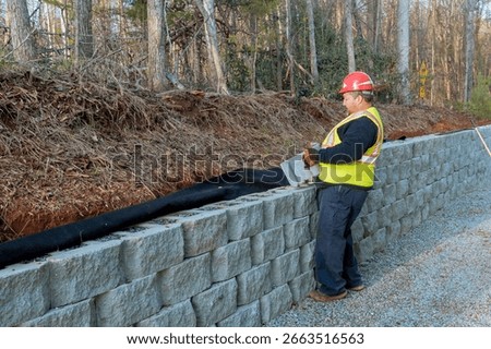 Worker arranges stone blocks for retaining wall by gravel road amid trees.