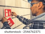 A worker attaches essential fire safety signs to a wall at a construction site, ensuring safety compliance during daylight hours.