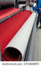 A worker in a blue jacket operates a machine that rolls a large red carpet onto a white backing material in a factory setting