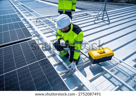 A worker in a bright yellow jacket and white helmet installs solar panels on a commercial building rooftop, using a power drill to secure mounting hardware