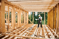 Worker Building Wooden Two-story House Near The Forest. Man In Work Clothes And Hard Hat Reviewing The Construction Plan. The Aim Is To Implement Modern, Environmentally-friendly Construction Methods.