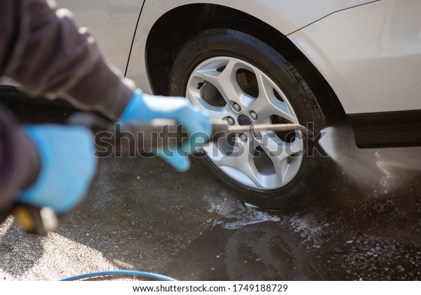 A worker in a car wash sprays a gray car with a\
strong jet of water. The worker wears blue rubber sneakers. Car\
wash. Copy Space.