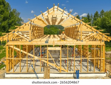 A worker carefully assembles the wooden framework of a house under a clear blue sky amidst picturesque greenery. The structure is in progress.