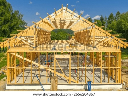 A worker carefully assembles the wooden framework of a house under a clear blue sky amidst picturesque greenery. The structure is in progress.