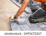 A worker carefully positions paving stones on a pathway, focusing on alignment and precision. Wearing gloves, the individual kneels on a gravel surface, surrounded by stacks of stone blocks. 