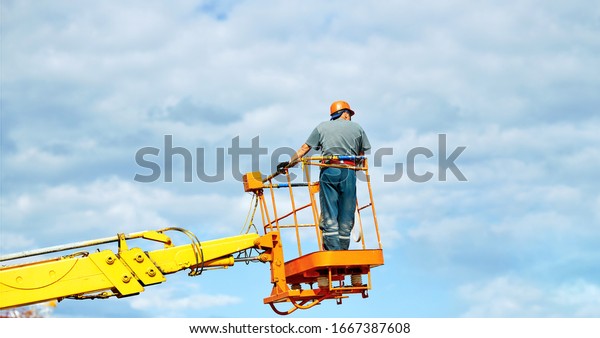 Worker in cherry picker bucket at height wearing\
hardhat and safety harness. Height construction worker against blue\
sky. Electrician standing in mobile crane bucket and rising to\
repair street wiring