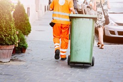 Worker Of Cleaning Company In Orange Uniform With A Green Garbage Bin