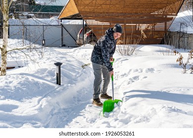 A Worker Cleans Snow From The Garden Paths With A Shovel On A Sunny Day. Garden Maintenance In Winter. Huge Snowdrifts After A Blizzard. The Yard Was Covered With Snow After A Heavy Snowfall.