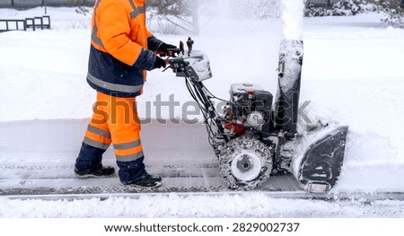 Worker clears snow with machine on sidewalk. Bright orange uniform contrasts with white snow. Snowblower forcefully ejects snow into air. Cold winter day with heavy snowfall