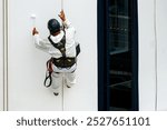 A worker with climber skills in mountaineering equipment paints the hull of a white cruise ship with a paint roller while hanging from a rope