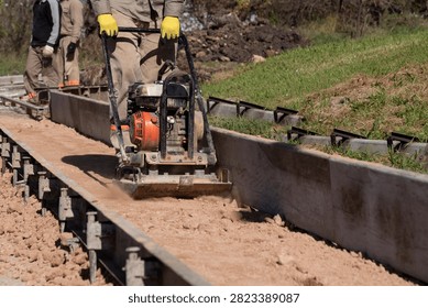 Worker compacting soil subbase with plate compactor between metal formwork for curb construction